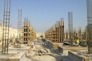 Construction site with multiple concrete and steel column frameworks extending horizontally. Reinforcement bars protrude from the columns, and wooden formwork is in place. A worker in a yellow safety vest is visible amidst the construction materials. Clear sky above.