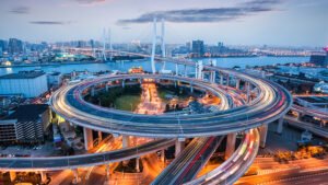 Aerial view of a complex, multi-level highway interchange with light trails from moving vehicles. The city skyline and a large suspension bridge are visible in the background, along with a river running parallel to the infrastructure. The evening sky is partly cloudy.