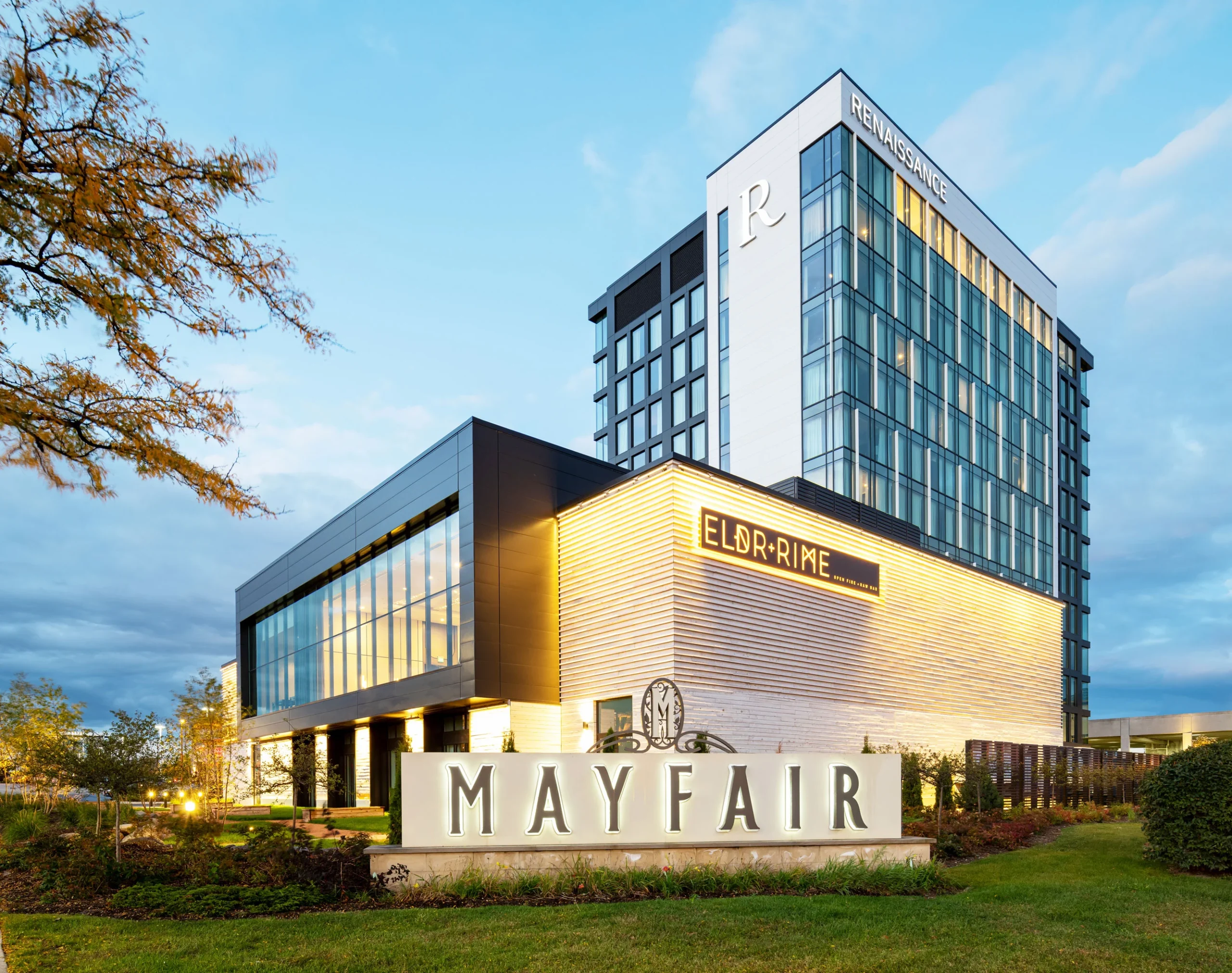 A modern hotel building with large glass windows and angular architecture, featuring signage for "Mayfair" and "Eldr+Rime". The building has a blend of dark and light tones with landscaped greenery in the foreground and a partially cloudy sky above.