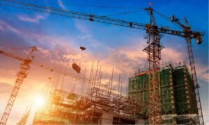 The image shows a construction site at sunset with several tall cranes towering over partially built structures covered in scaffolding. The sky is painted with hues of orange, pink, and blue, with the sun partially visible on the left side, casting a warm glow over the scene.