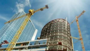 Construction site with a partially built skyscraper and two large yellow cranes set against a blue sky with bright sunlight. Adjacent to the construction, a completed glass-faced building stands. The scene depicts modern urban development.