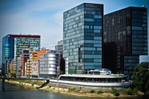 A cityscape displaying modern architecture along a river. Two tall, dark glass buildings bear the "Hyatt Regency" name. Surrounding structures showcase various designs, including a colorful, patterned building on the left. A waterfront promenade lines the riverbank.