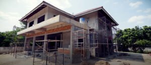 A large two-story house under construction, with scaffolding surrounding the building. The house has a red-tiled roof, unfinished concrete walls, and exposed brickwork. Construction materials and tools are scattered around the site, and the sky is clear and sunny.