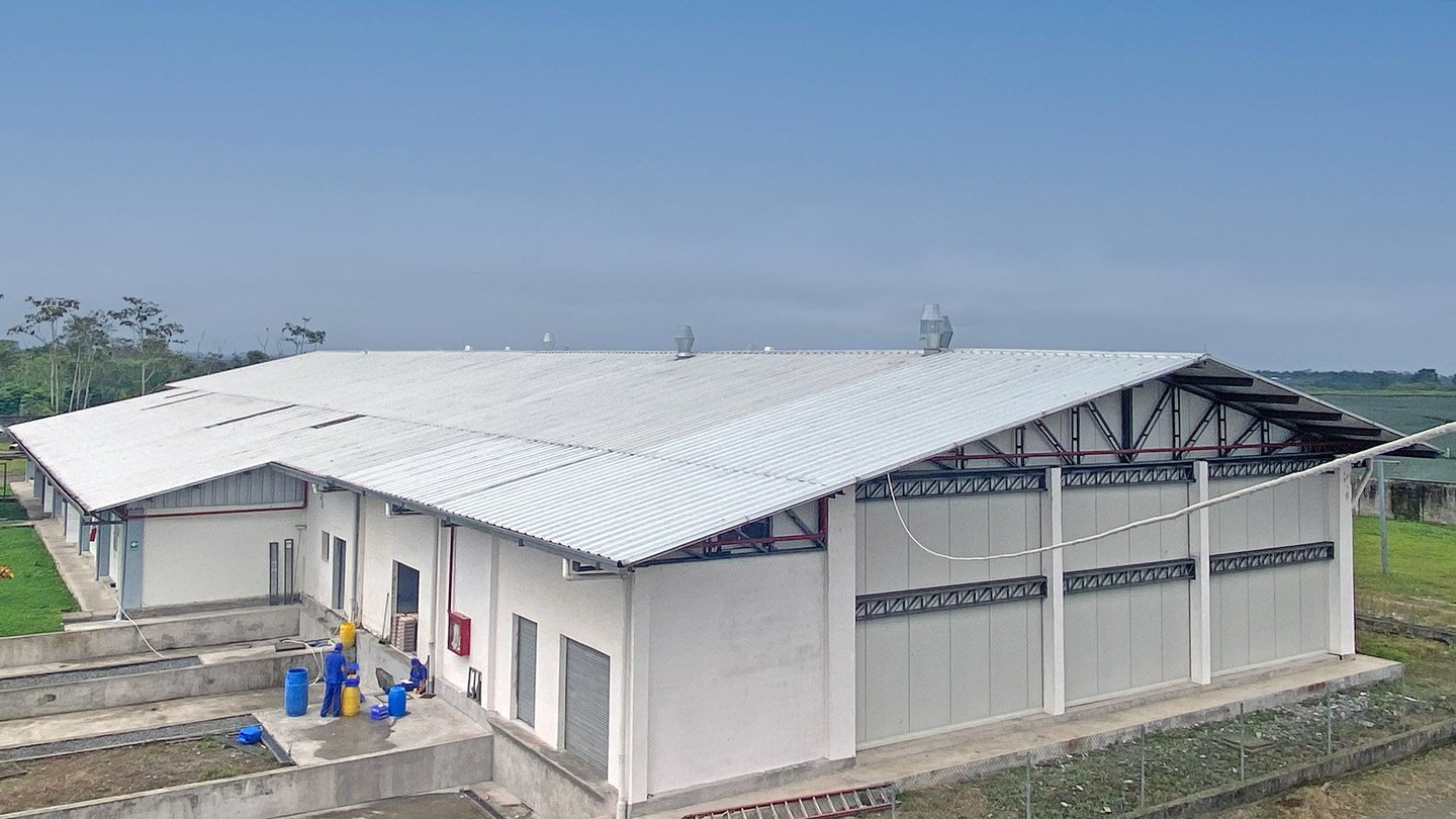 A large, white industrial building with a metal roof sits in a spacious, grassy area. Numerous windows line its walls, and several blue barrels and equipment are visible outside. The sky above is clear, enhancing the overall brightness of the scene.