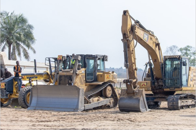 Construction site with a bulldozer and an excavator working on a sandy area. Several workers in safety gear stand nearby, observing or assisting the operation. A backdrop of trees and equipment trailers can be seen in the distance.